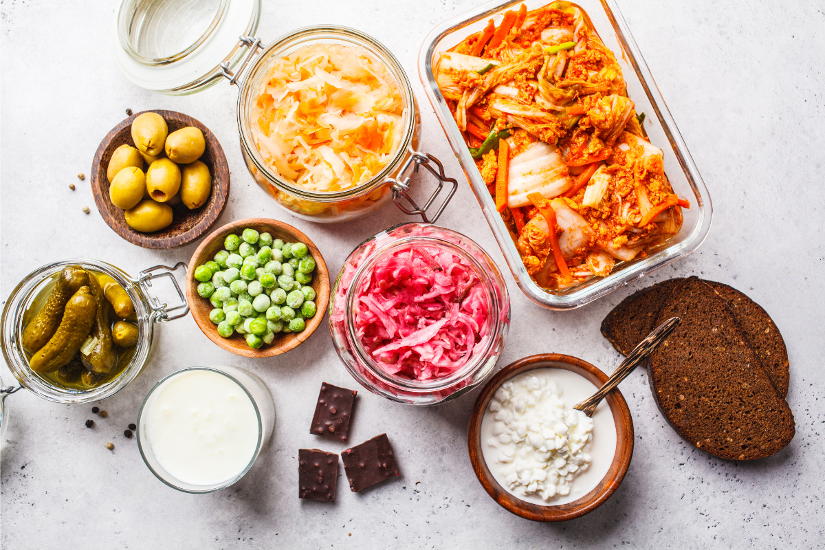 A variety of fermented foods including kimchi, sauerkraut, pickles, olives, yoghurt, cottage cheese, and dark chocolate displayed on a table.