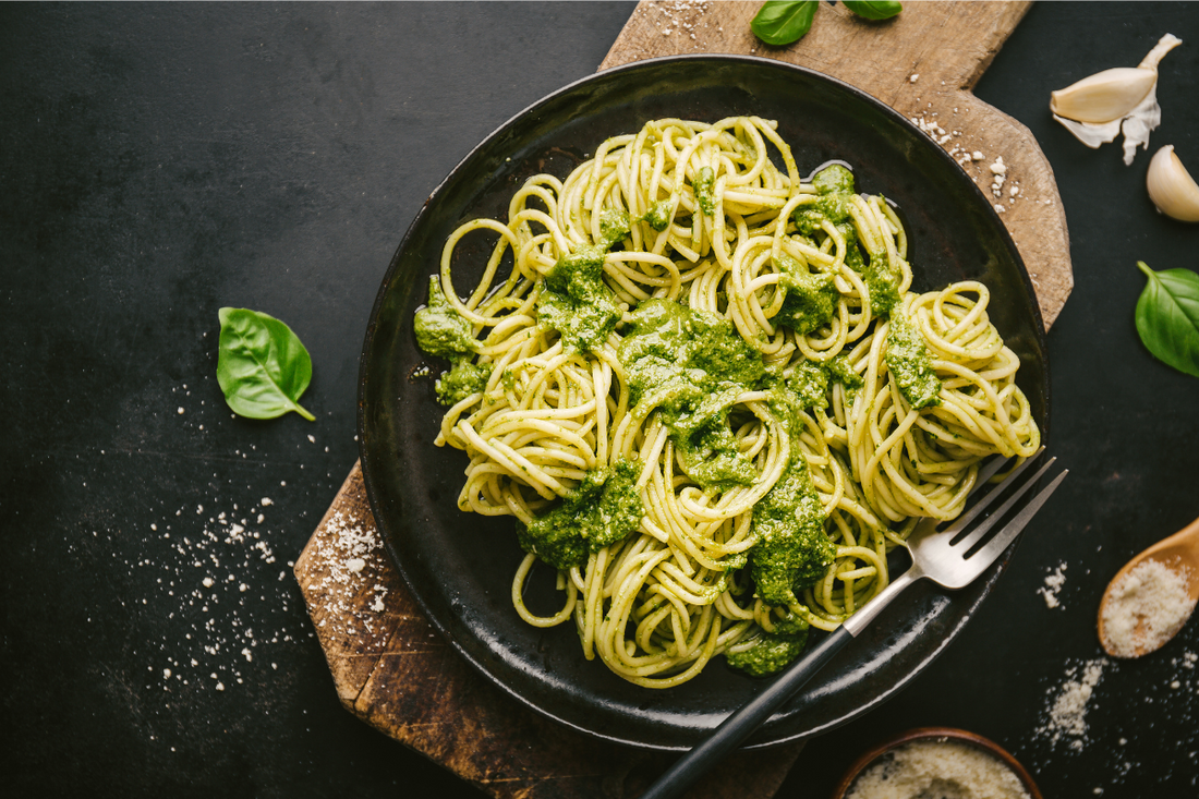 A plate of spaghetti coated in green broccoli pesto sauce, garnished with fresh basil and Parmesan cheese on a dark rustic background.