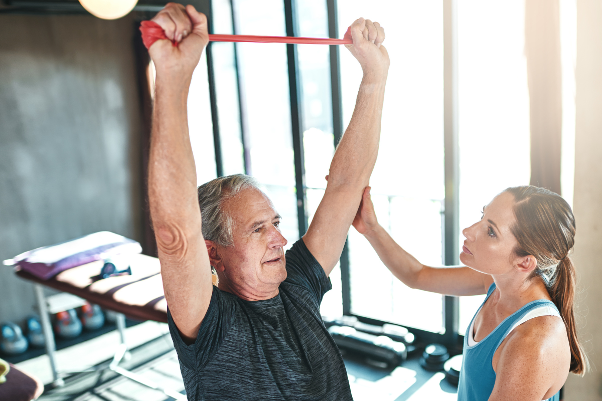 A senior man lifting a resistance band over his head with the assistance of a female trainer in a gym setting.