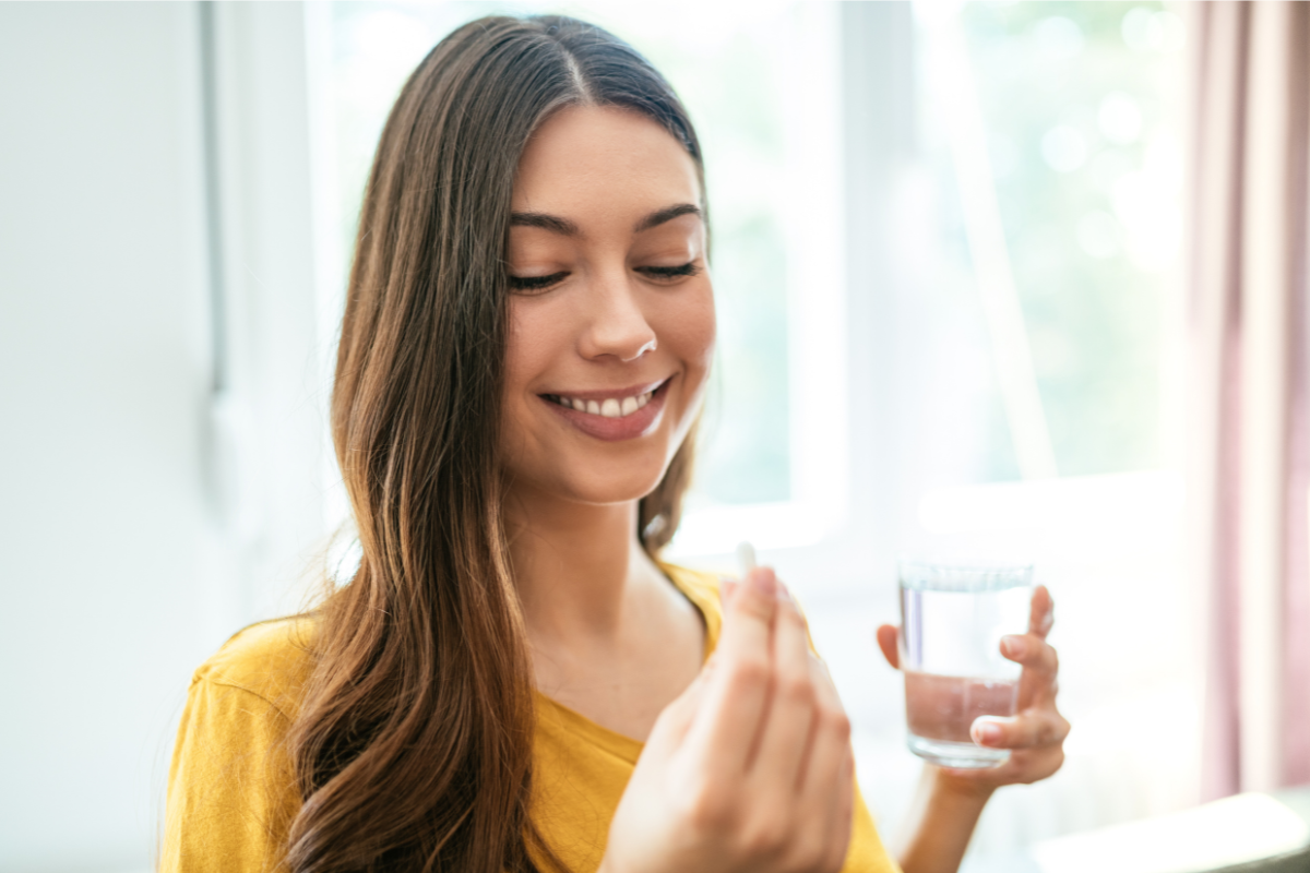 A smiling woman holding a glass of water and a supplement capsule, preparing to take it.