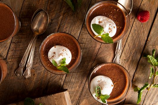 Glasses of dark chocolate avocado mousse topped with cream and mint, arranged on a rustic wooden table with vintage spoons.