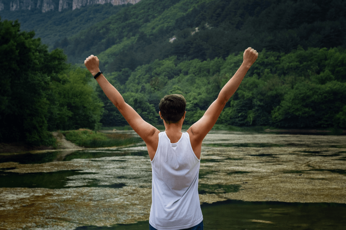 Man in a white tank top standing near a lake with arms raised.