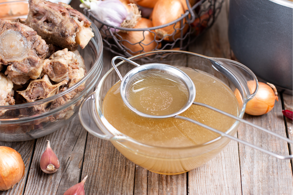 Glass bowl of homemade bone broth with strainer on a wooden surface.