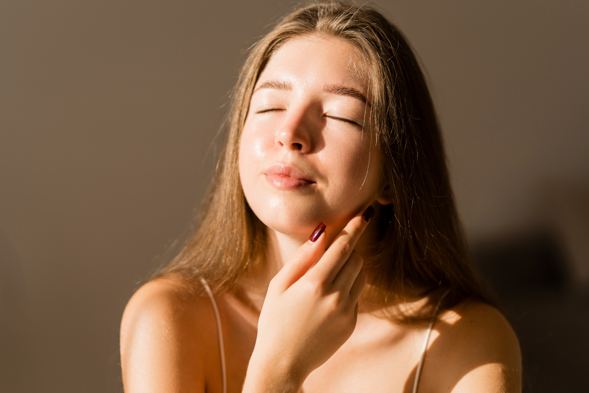Woman with eyes closed, gently touching her face in soft natural light.