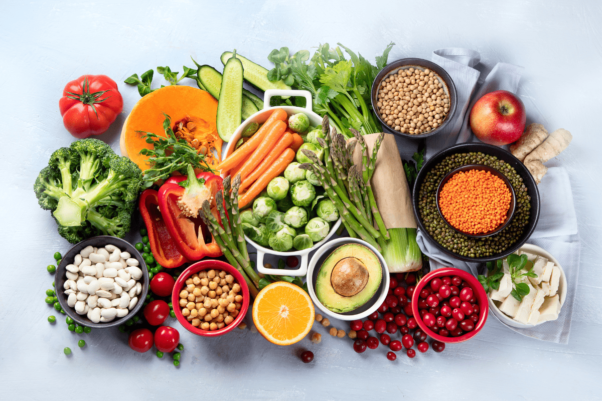 Assortment of fresh vegetables, fruits, legumes, and grains arranged on a light background.