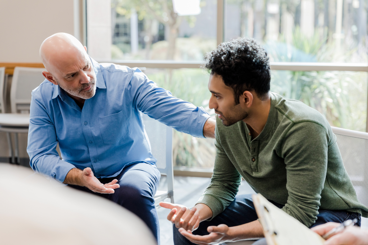 A bald man in a blue shirt listens attentively to a younger man in a green sweater, offering a comforting hand on his shoulder.