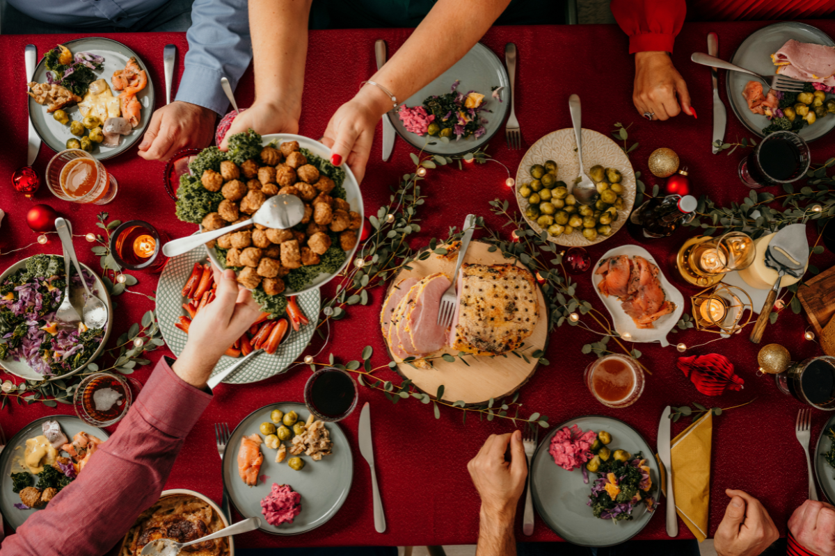 Festive holiday table with ham, appetizers, and guests reaching for food.