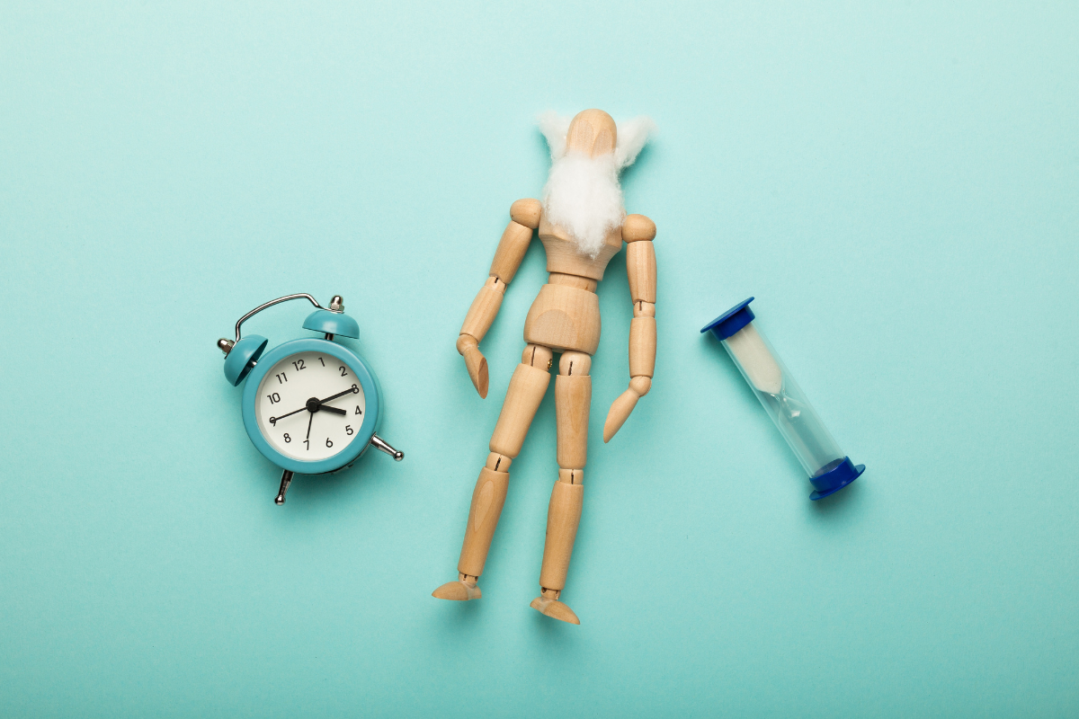 A wooden mannequin with a white beard lies on a blue background, flanked by a small alarm clock and an hourglass.