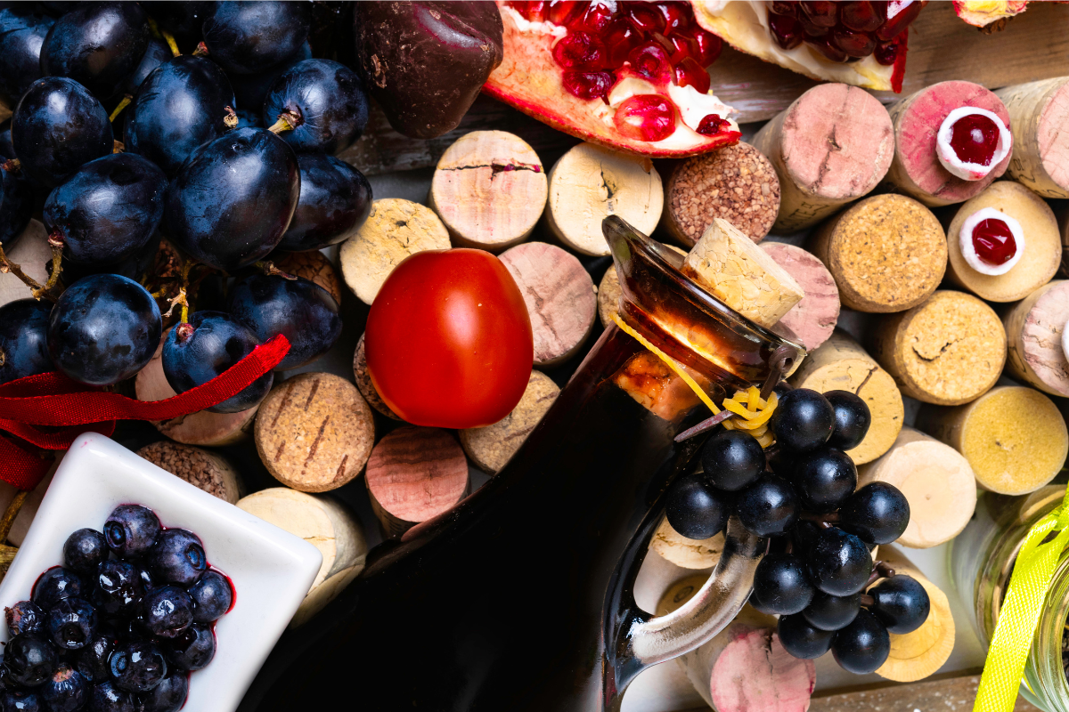 A variety of fruits and vegetables, including grapes, blueberries, pomegranate, and a tomato, arranged with wine corks and a bottle of wine.