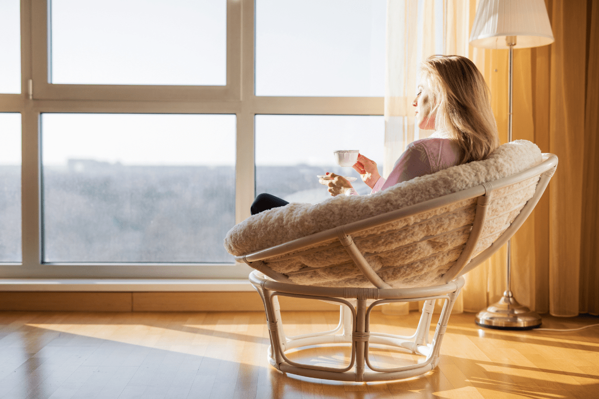 Woman sitting in a cosy chair by a large window, enjoying a cup of tea in a sunlit room.