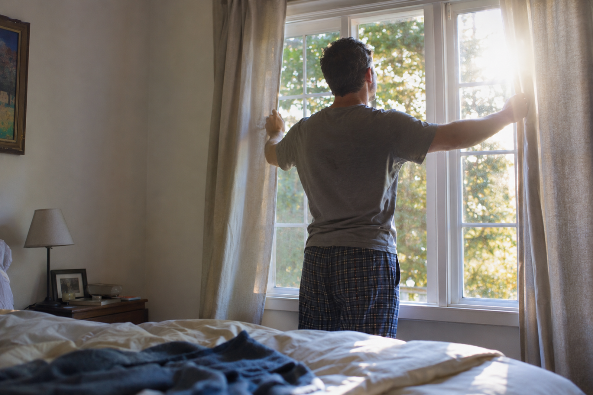 Middle-aged man opening curtains in a sunlit bedroom.