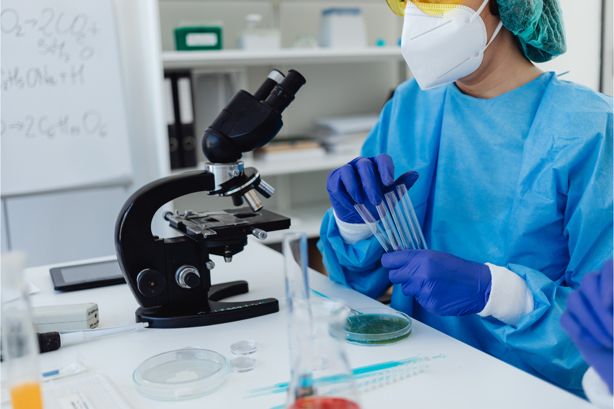 Scientist in protective gear analyzing samples with a microscope and test tubes in a laboratory.