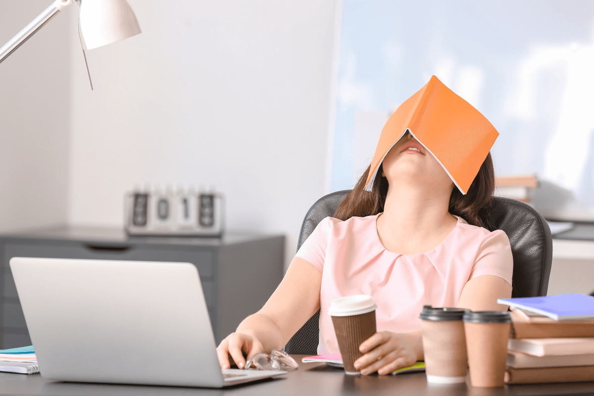 A tired woman at her desk with a book covering her face, holding a coffee cup, surrounded by work materials.