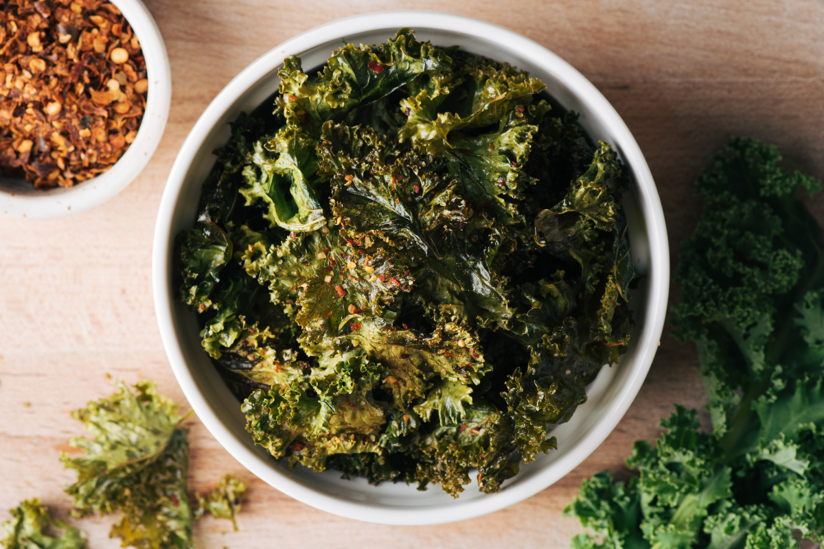 A bowl of crispy, seasoned kale chips with a side dish of crushed red pepper flakes.