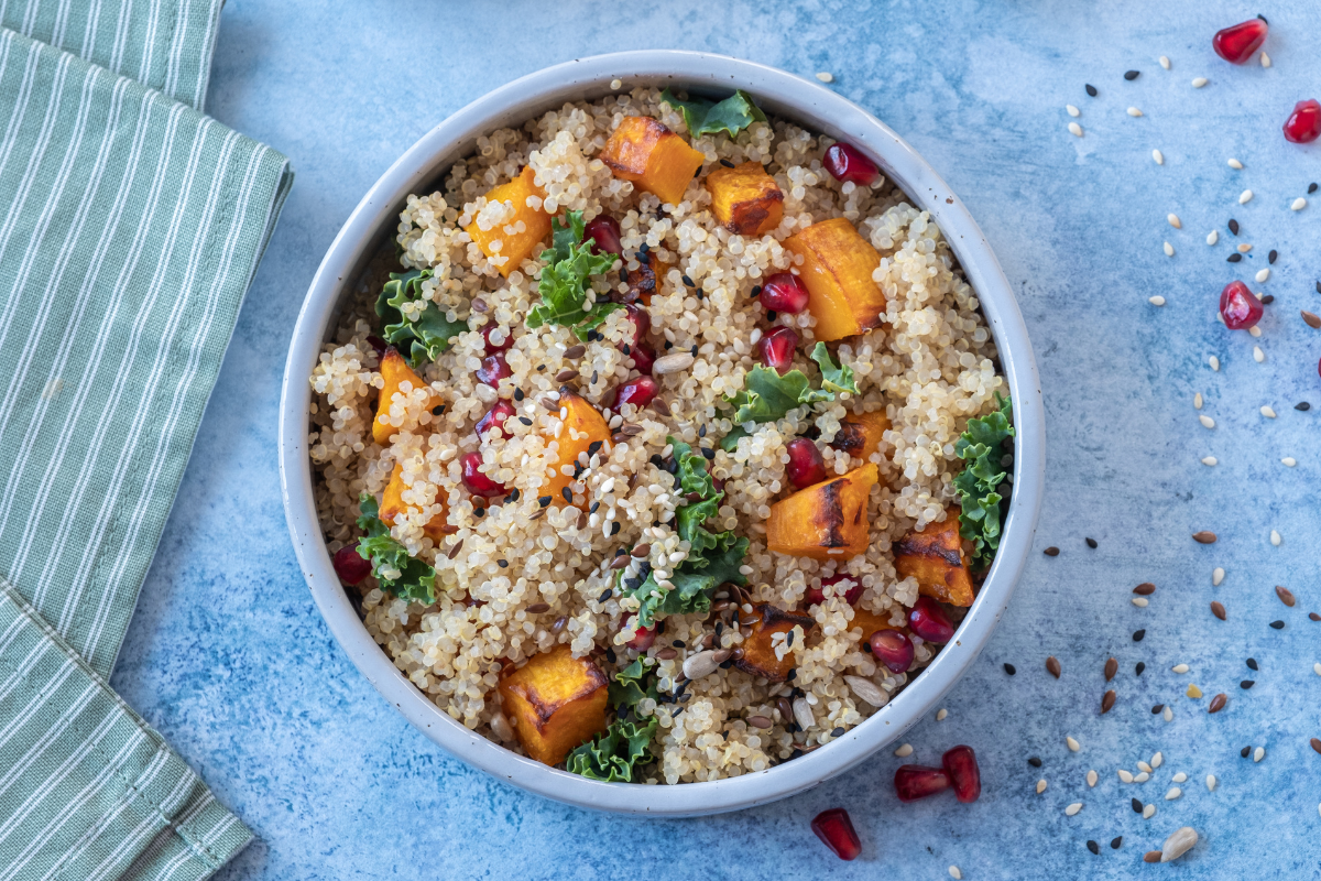 A vibrant bowl of quinoa mixed with roasted sweet potatoes, pomegranate seeds, fresh kale, and sprinkled with seeds, sitting on a blue background with scattered ingredients nearby.