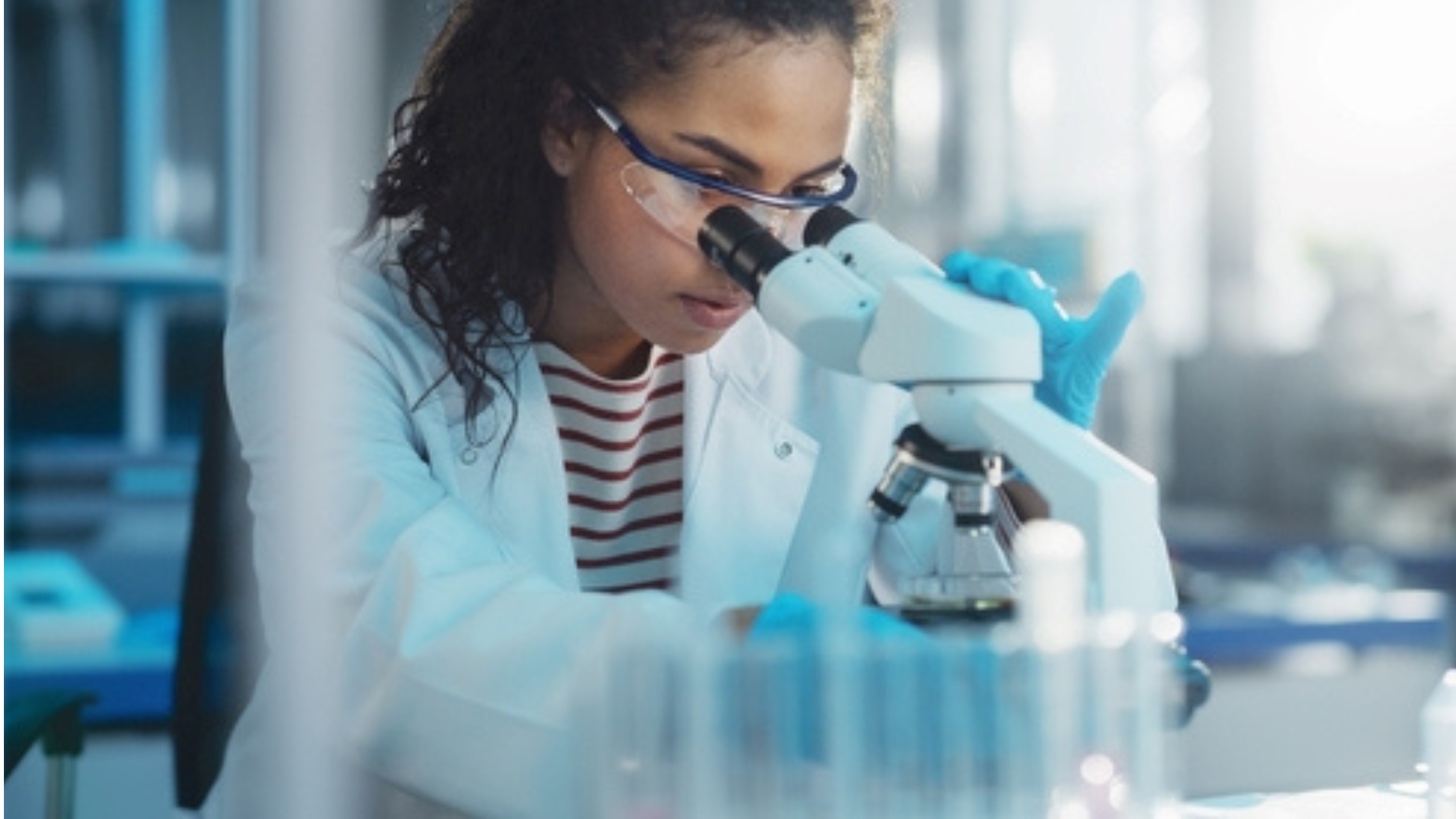 A woman wearing a lab coat is seen looking into a microscope in a laboratory setting.