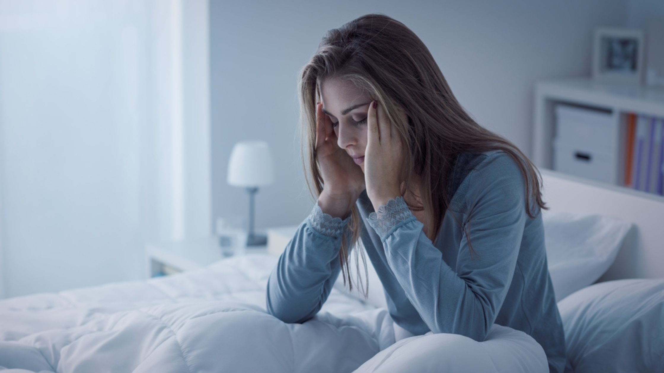 A woman sitting on a bed, holding her temples with a distressed expression, indicating difficulty sleeping