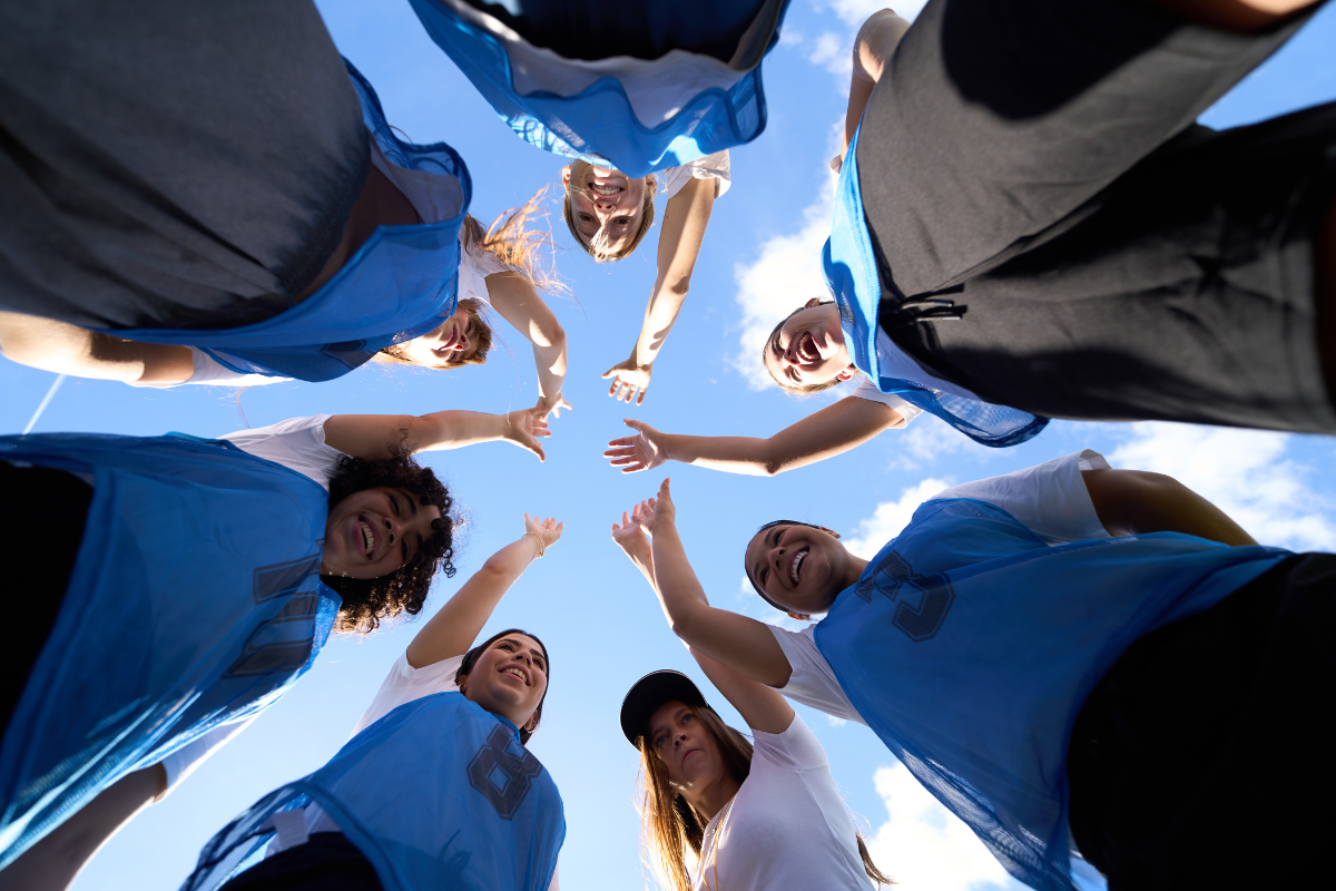 Group of people in circle looking down at camera with hands reaching in together.