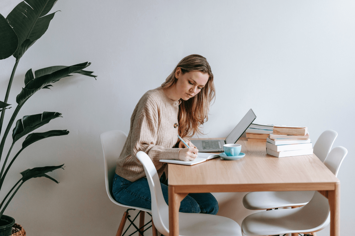 Young woman writing in a notebook at a table with a laptop and books.